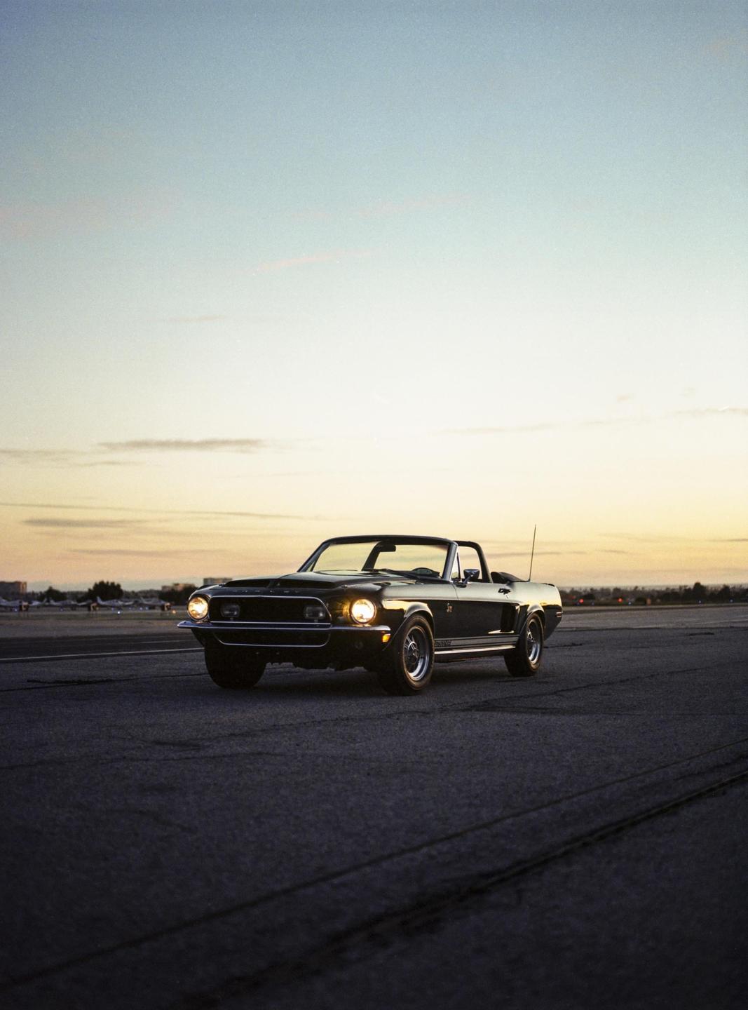A black convertible Mustang is parked on an airfield at dusk.