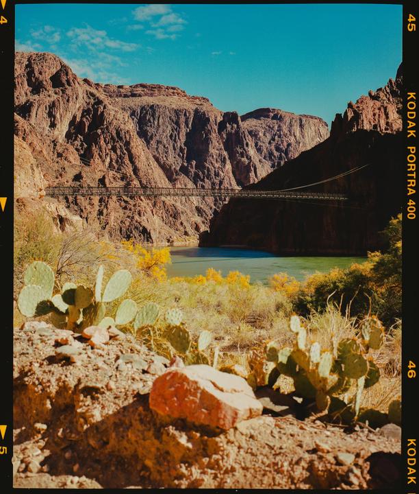 A bridge spans a river between two large rock formations, with cacti and rocks in the foreground.