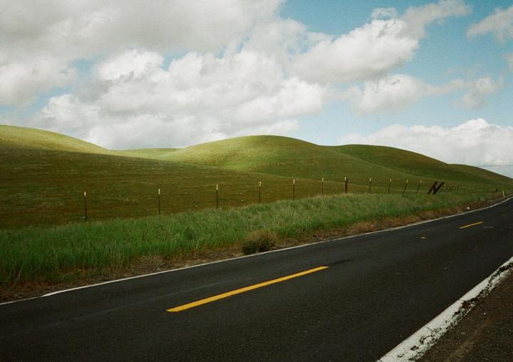 A road with a yellow dividing line runs alongside green rolling hills under a cloudy sky.