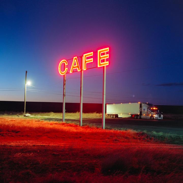 A neon sign for a cafe stands beside a road with a truck parked nearby at night.