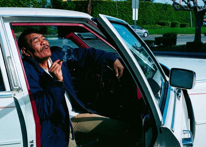 A man with a mustache and sunglasses is sitting in a white car with the door open, smoking a cigar.