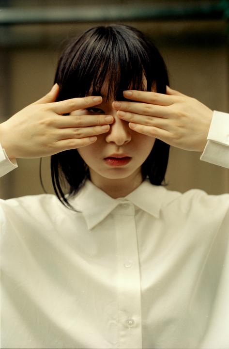 A woman with dark hair covers her eyes with her hands while wearing a white collared shirt.