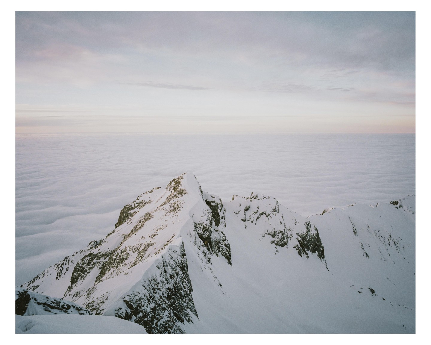 A snow-covered mountain peak rises above a sea of clouds under a pale sky.
