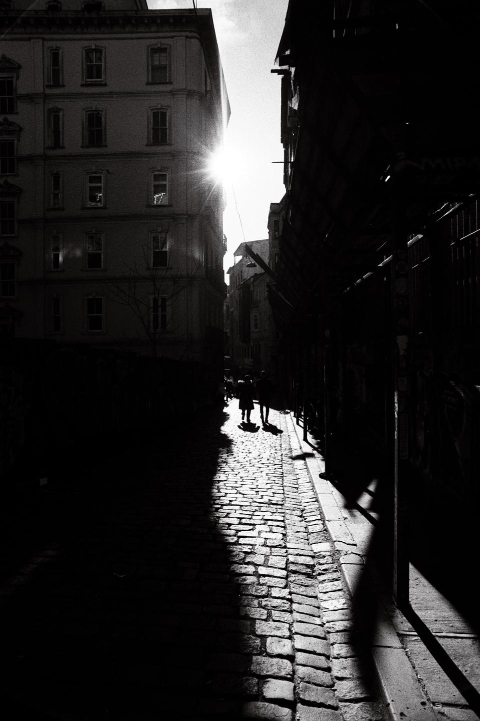 Two people walk on a sunlit cobblestone street between tall buildings, with one side of the street in deep shadow.