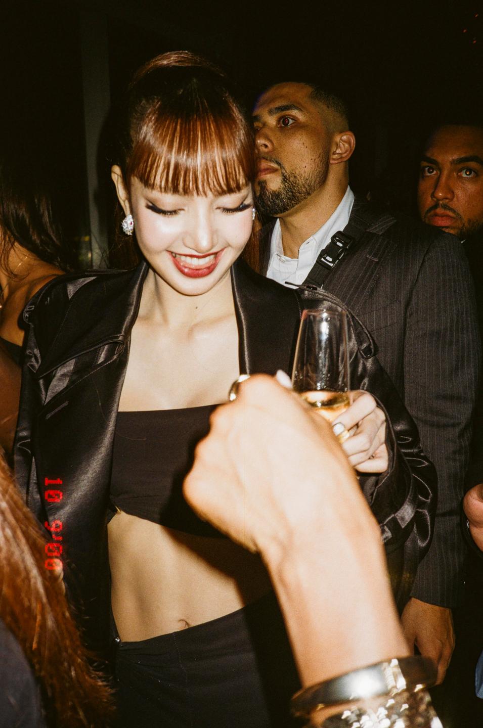 A woman with bangs and a black top smiles while holding a drink in a crowded, dimly lit room.