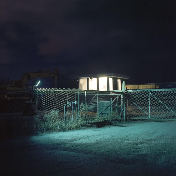 A brightly lit security booth stands behind a chain-link fence at night, with an excavator visible in the background.