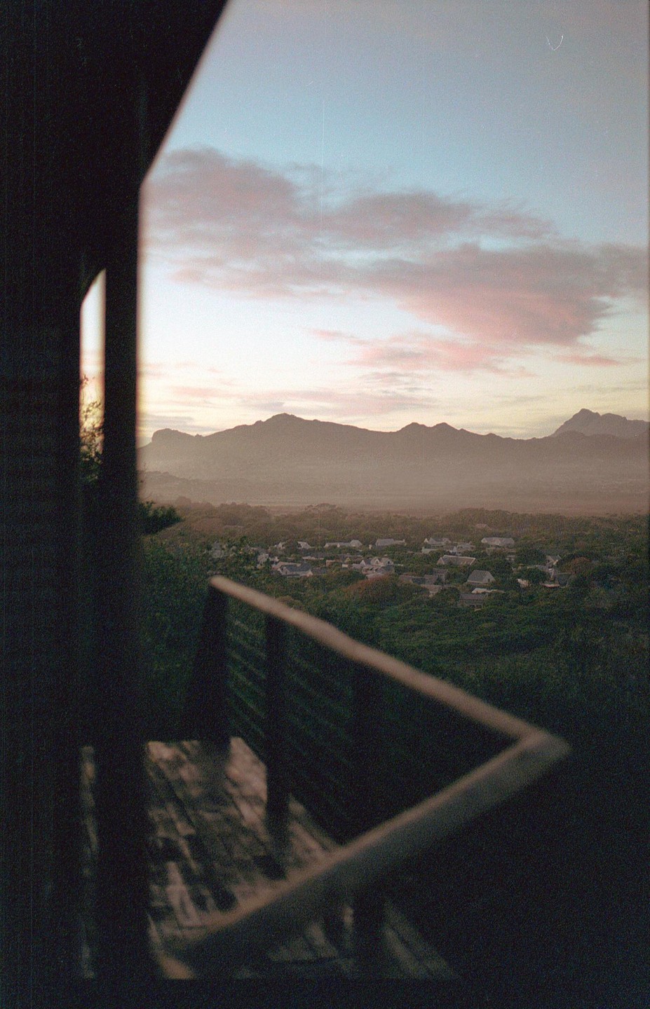 A view from a balcony shows a town nestled in a valley with mountains in the distance under a pastel sky.