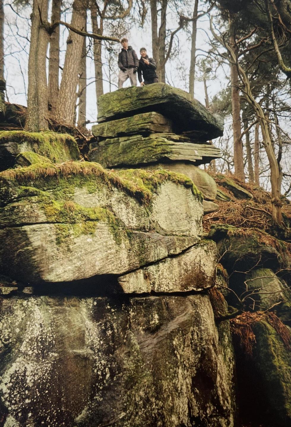 Two men stand on a mossy rock formation in a forest.