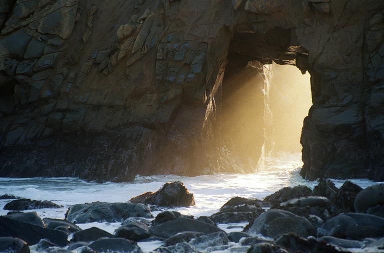 Sunlight streams through a natural rock archway over a rocky, foamy ocean.