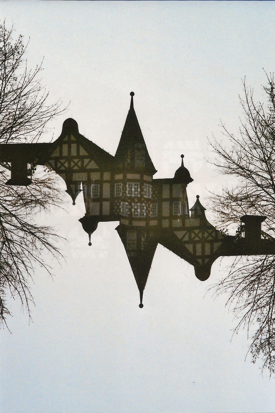 A half-timbered building with turrets is reflected in water, framed by bare tree branches.