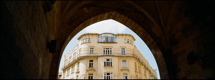 A large, light-colored building with many windows is framed by a dark stone archway.