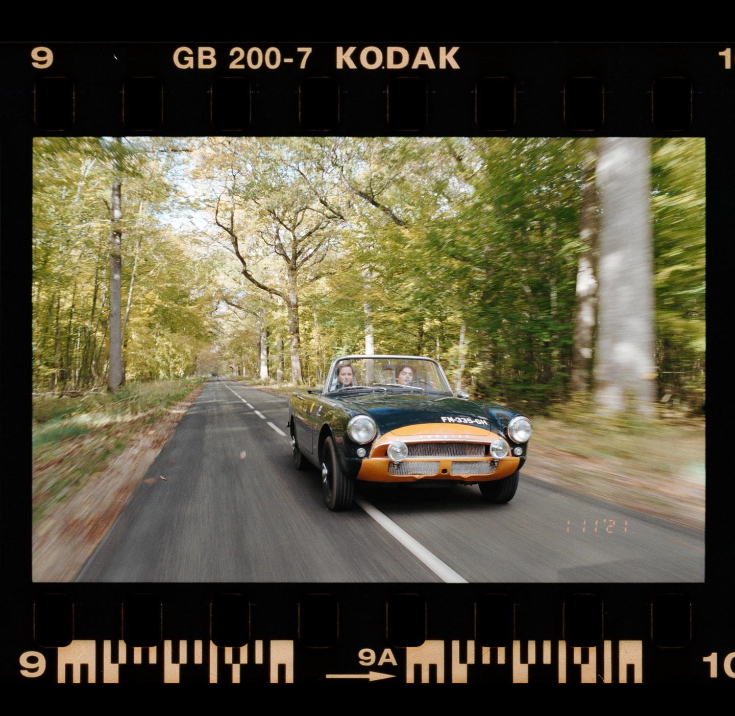 A convertible car with two women drives down a road lined with autumn trees.