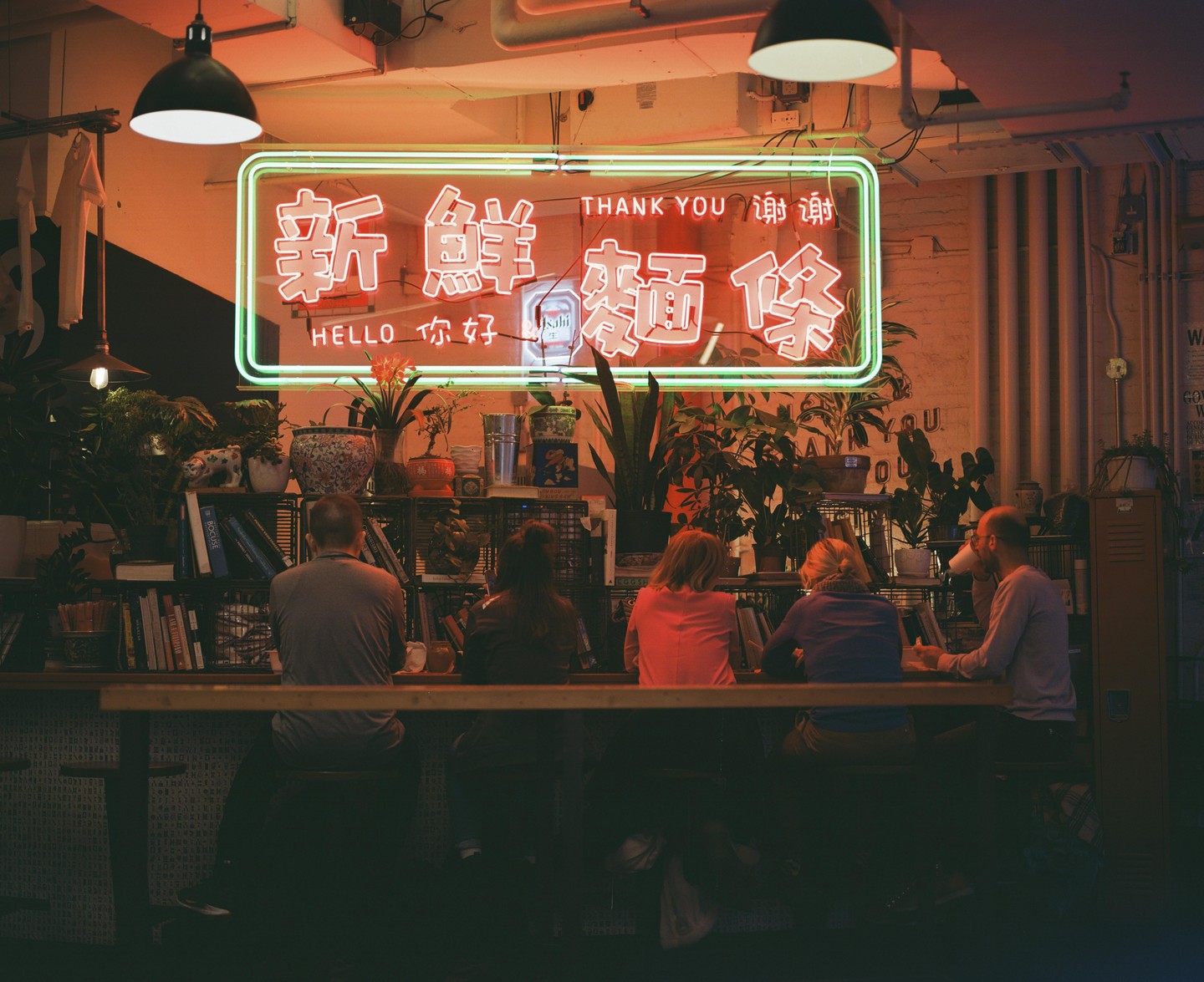 A group of people sit at a bar beneath a neon sign with Chinese characters and English words.