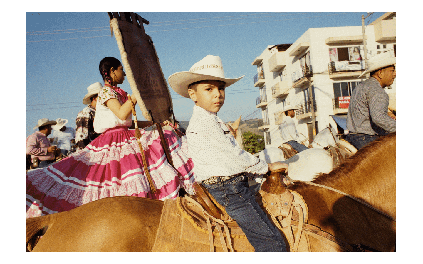 A boy in a cowboy hat sits on a horse next to a woman in a pink dress, with other people on horseback and buildings in the background.