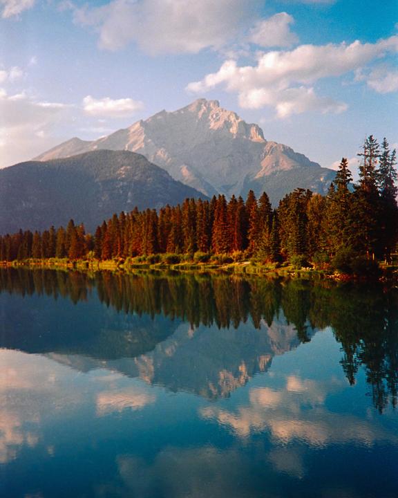 A mountain range and a forest are reflected in a calm lake under a blue sky with clouds.