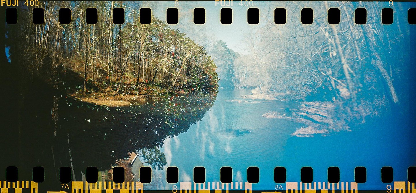 A double exposure shows a forest with a river and a bright blue sky with clouds.