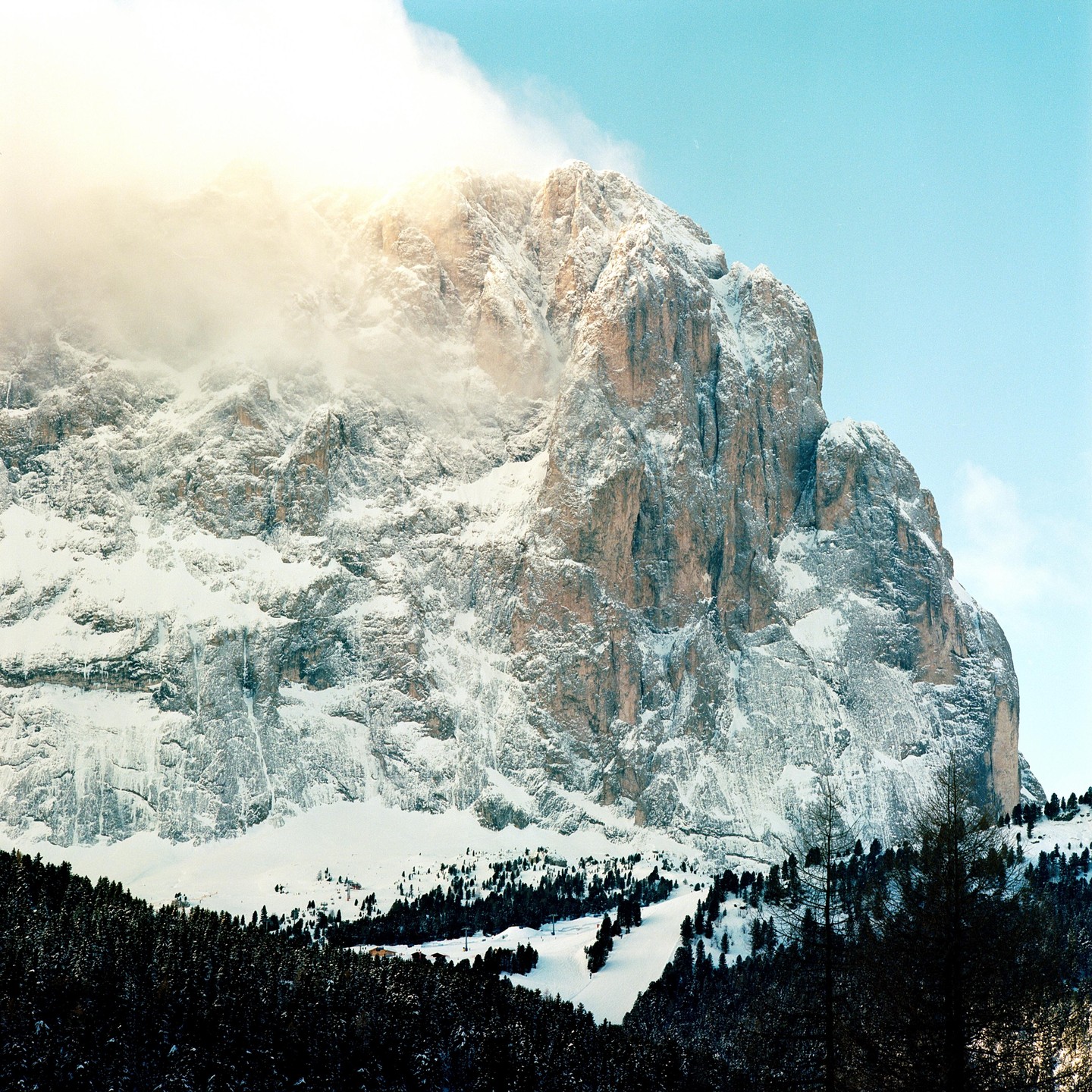 A snow-covered mountain with a forest at its base is partially obscured by clouds and bright light.