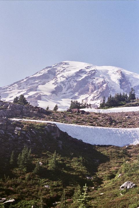 A snow-capped mountain rises above a rocky, forested slope with patches of snow.