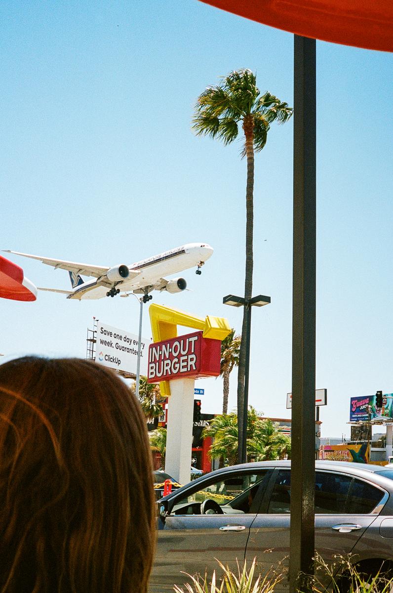 A large airplane flies low over a street with an In-N-Out Burger sign and palm trees.