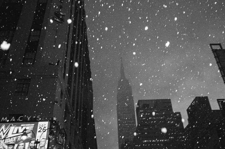Snow falls over tall buildings in a city at night.