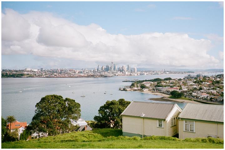 A panoramic view of Auckland, New Zealand, shows the city skyline across a body of water with boats, and houses in the foreground.