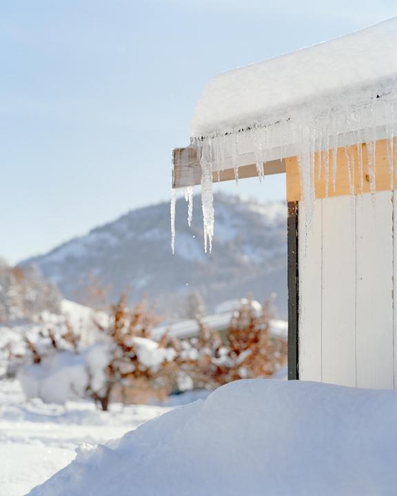 Icicles hang from the snow-covered roof of a white building with a snowy mountain in the background.