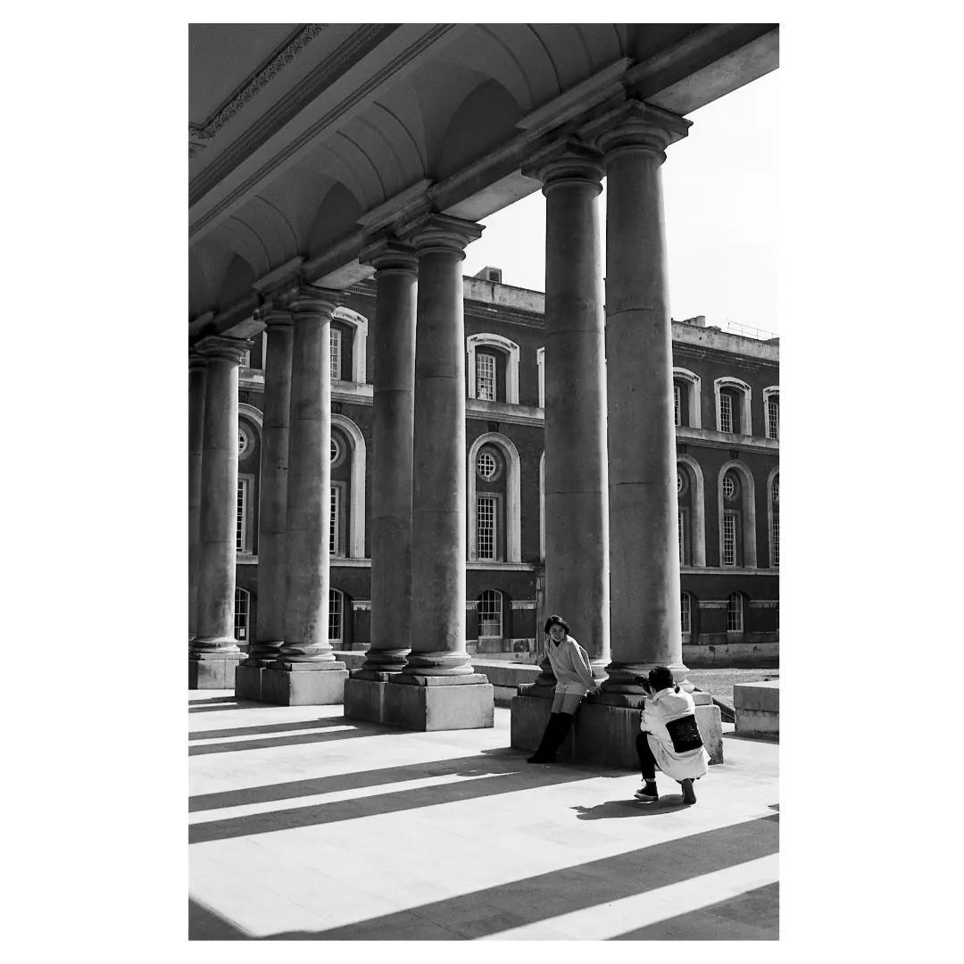 Two women are visible under a colonnade with a large building in the background.