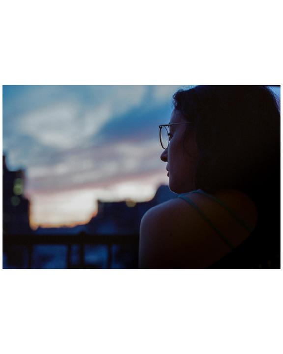 A woman with glasses looks out at a sunset over a city.