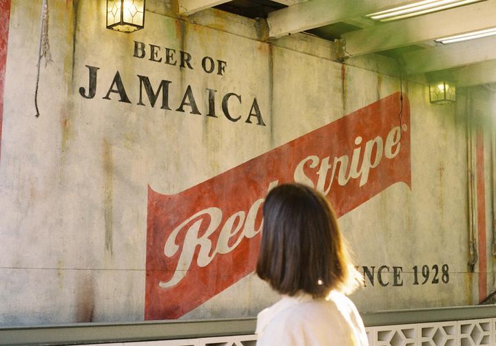 A woman stands with her back to the camera, looking at a wall with a large "Beer of Jamaica Red Stripe Since 1928" advertisement.