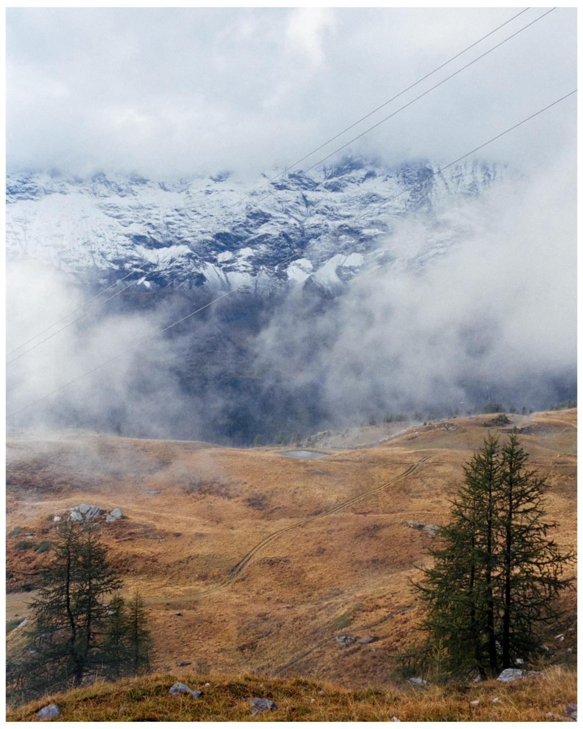 A snow-capped mountain range is partially obscured by clouds above a golden-brown hillside with scattered trees and power lines.