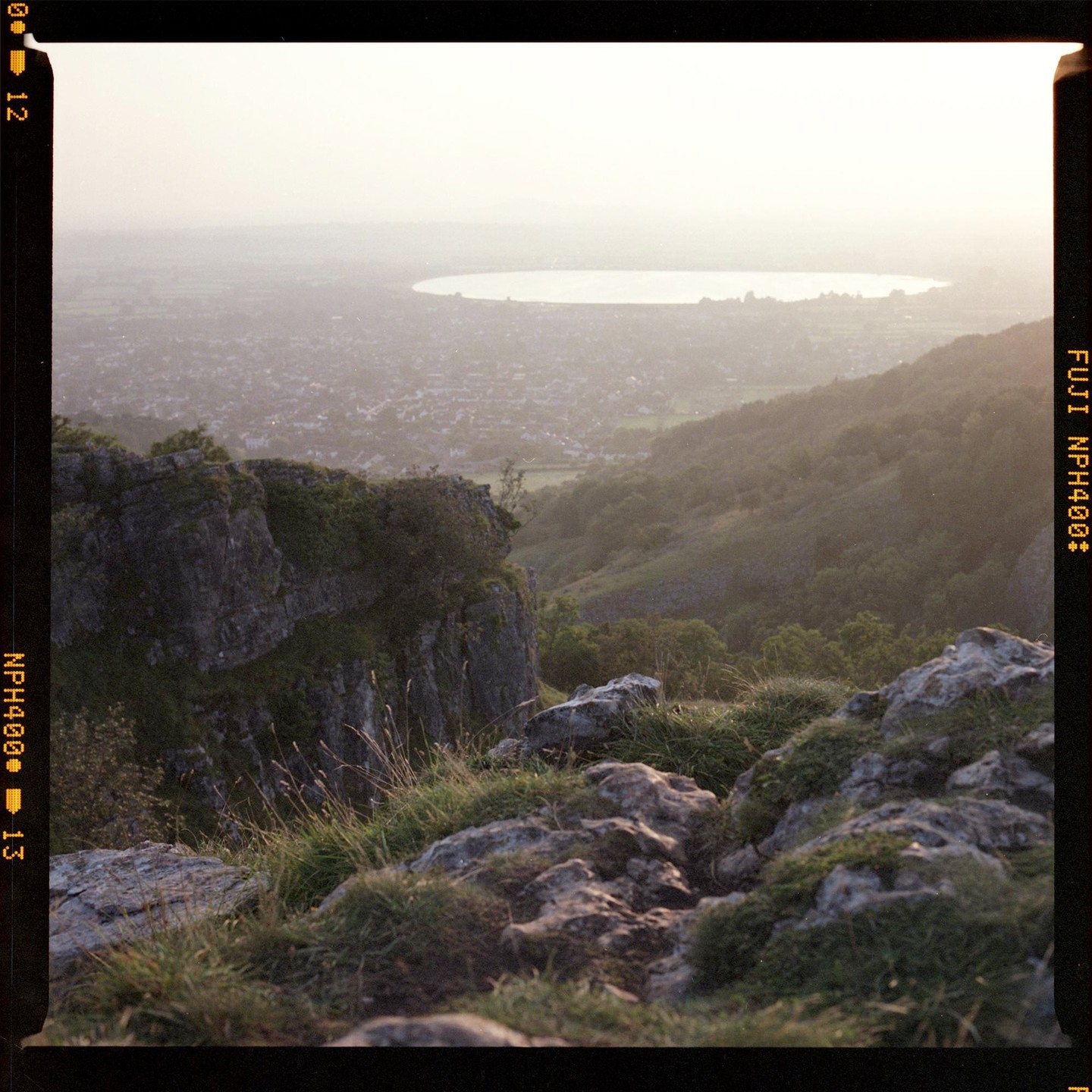 A hazy landscape shows a valley with a town and a lake, viewed from a rocky, grassy cliff.