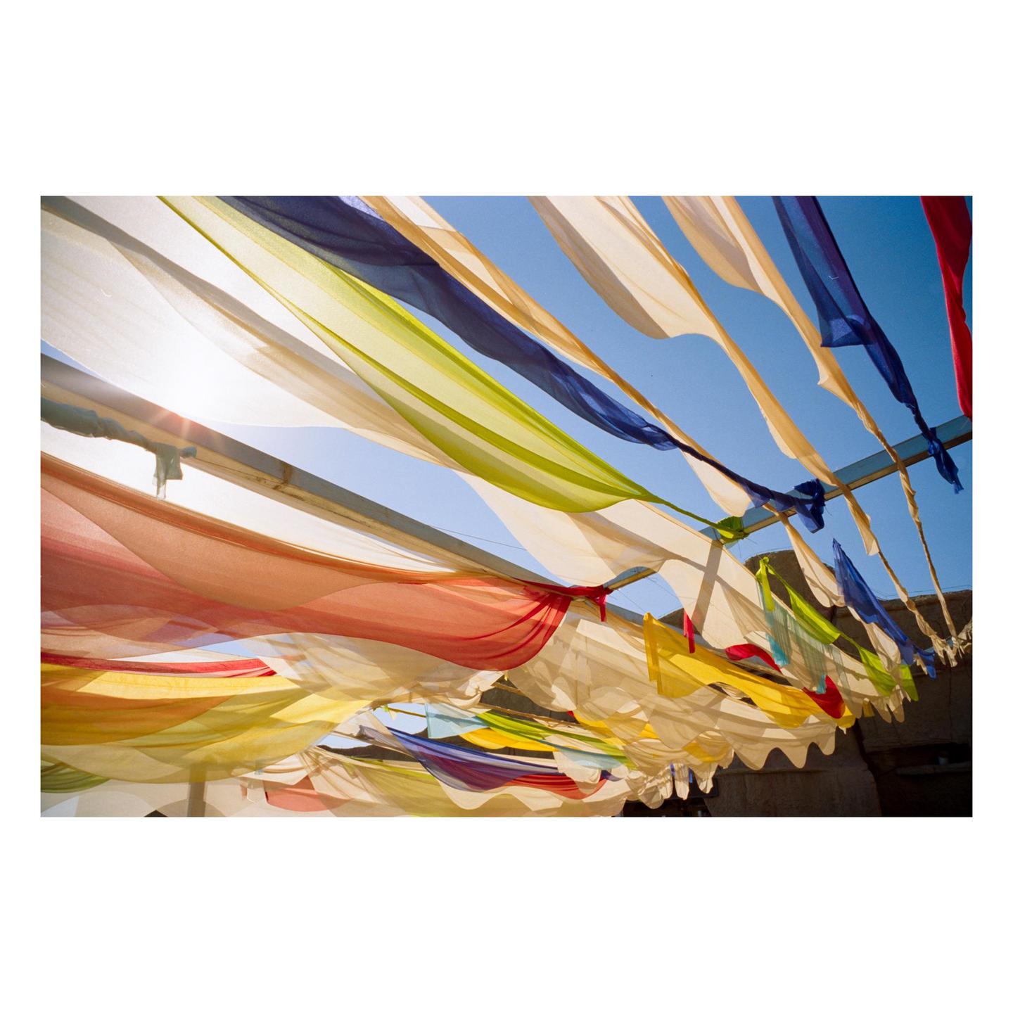 Colorful fabric strips are strung across a blue sky, with sunlight shining through them.