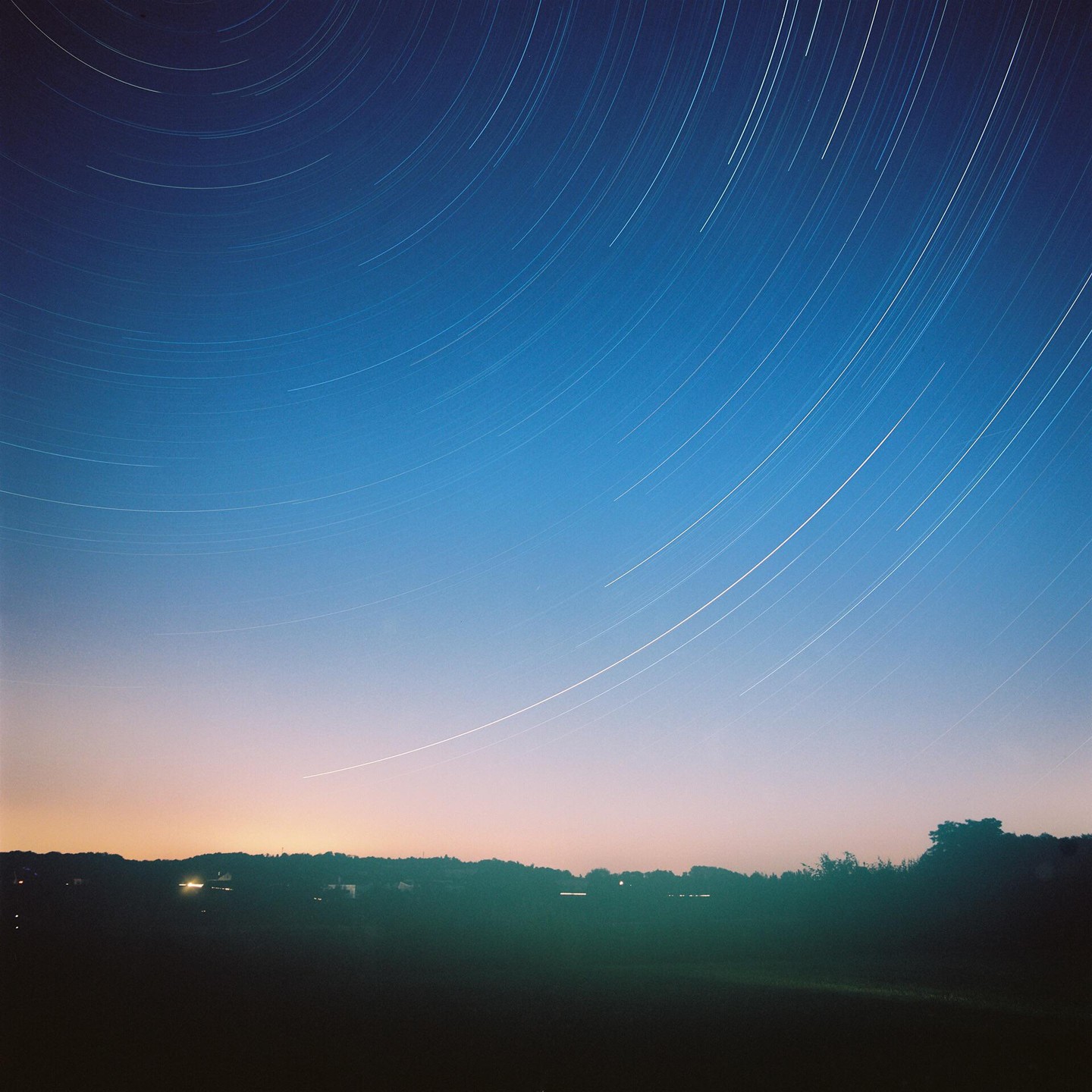 The night sky is filled with star trails above a dark landscape with distant lights.