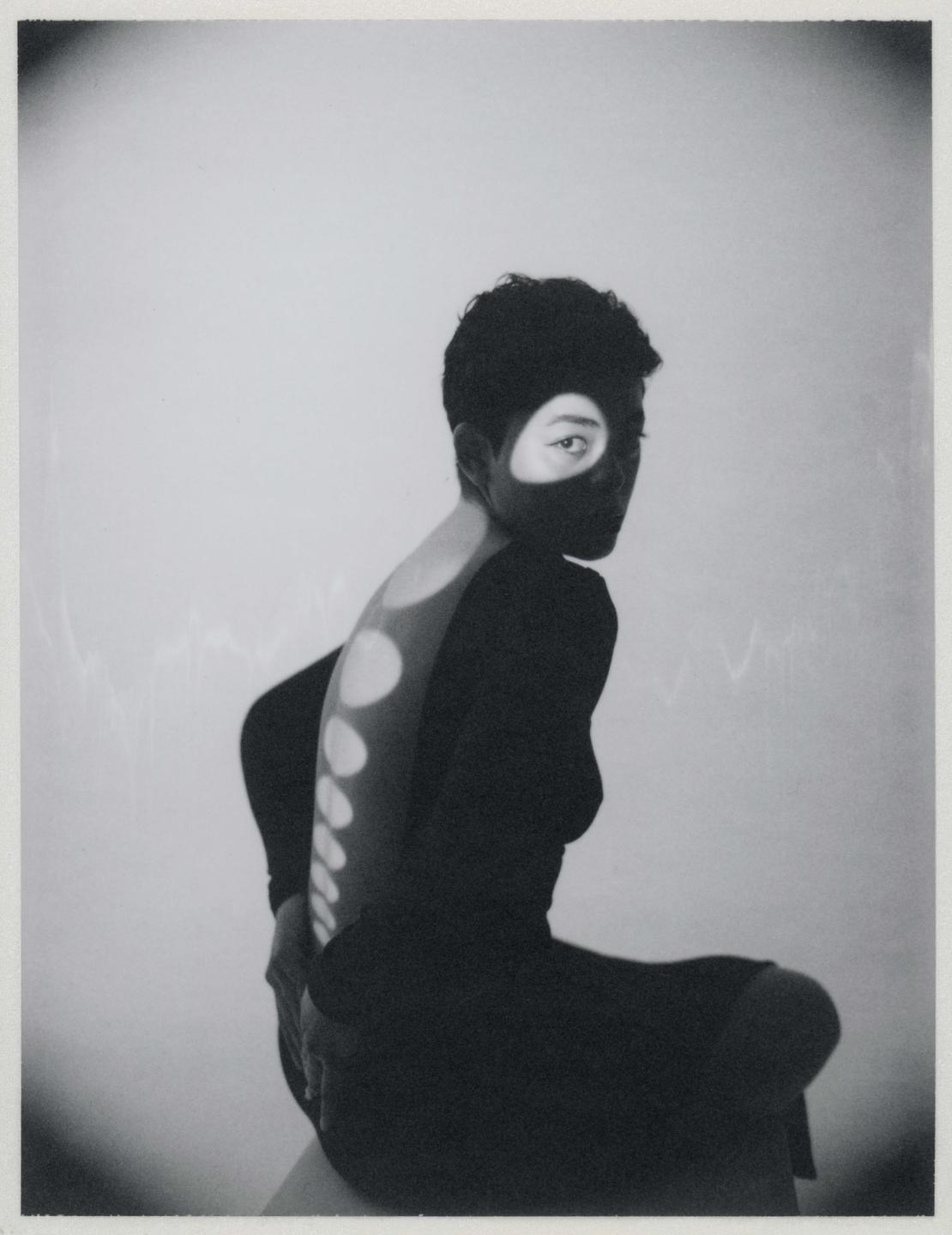 A woman with short hair sits with her back to the viewer, looking over her shoulder, with light projected onto her face and back.
