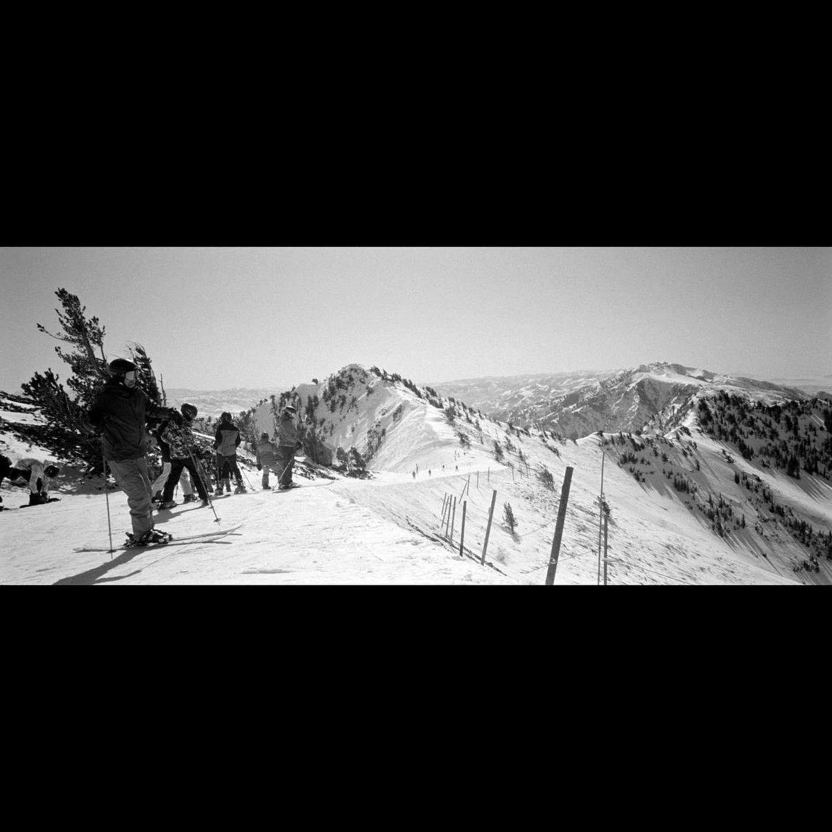 A group of skiers stands on a snowy mountain ridge with a vast mountain range in the background.
