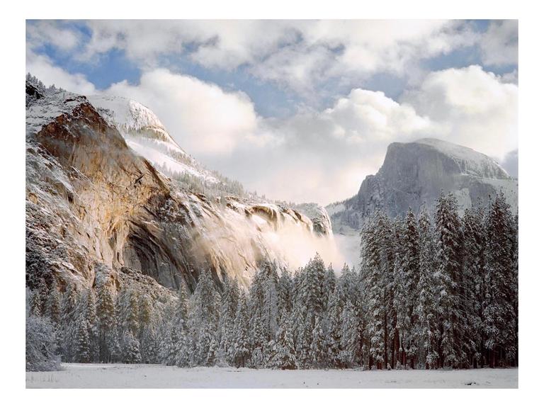A snow-covered valley with pine trees and mountains is illuminated by morning light under a cloudy sky.
