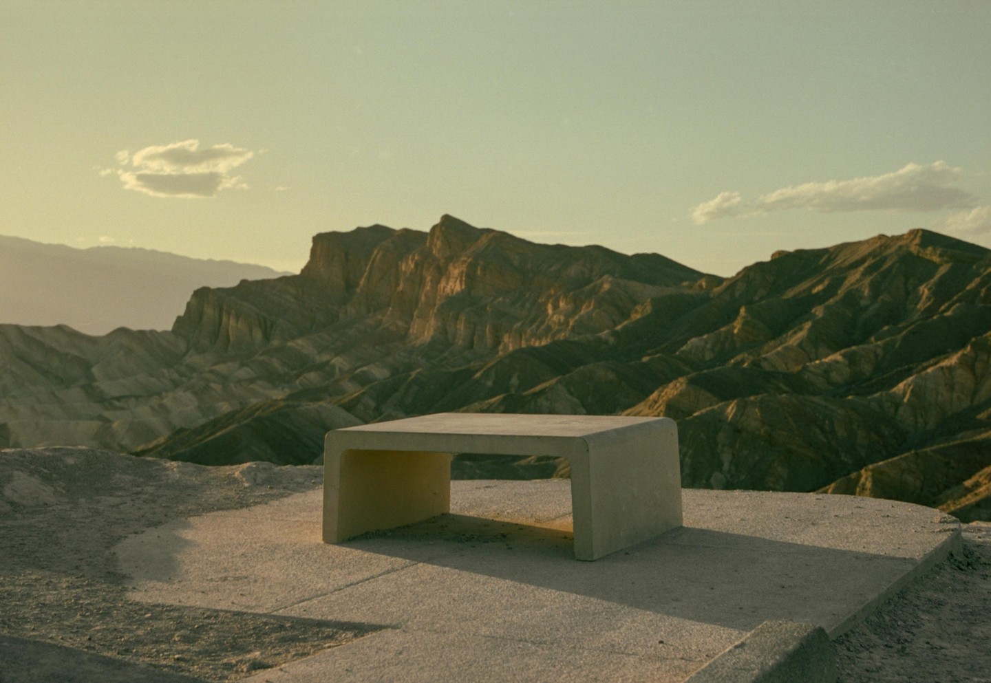 A concrete bench sits on a paved surface overlooking a vast desert mountain range under a clear sky.
