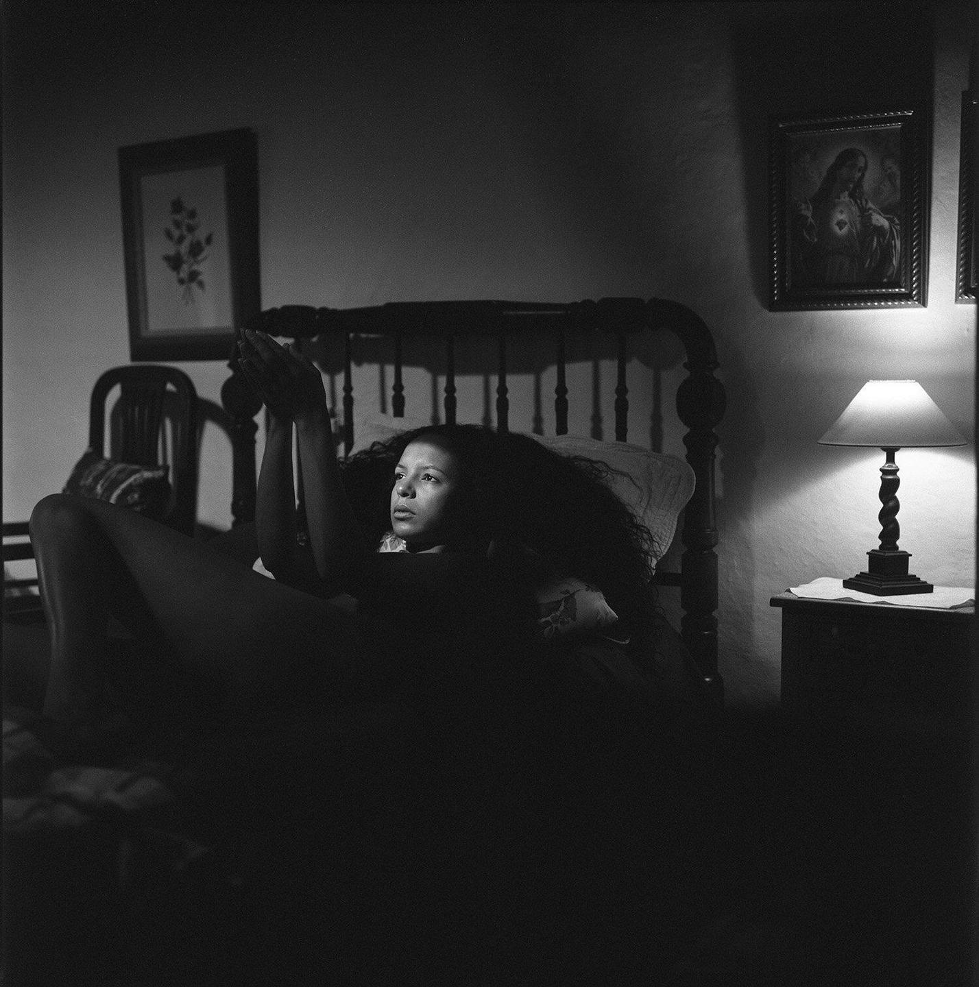 A woman with long, curly hair lies in bed with her hands raised, illuminated by a lamp on a bedside table.