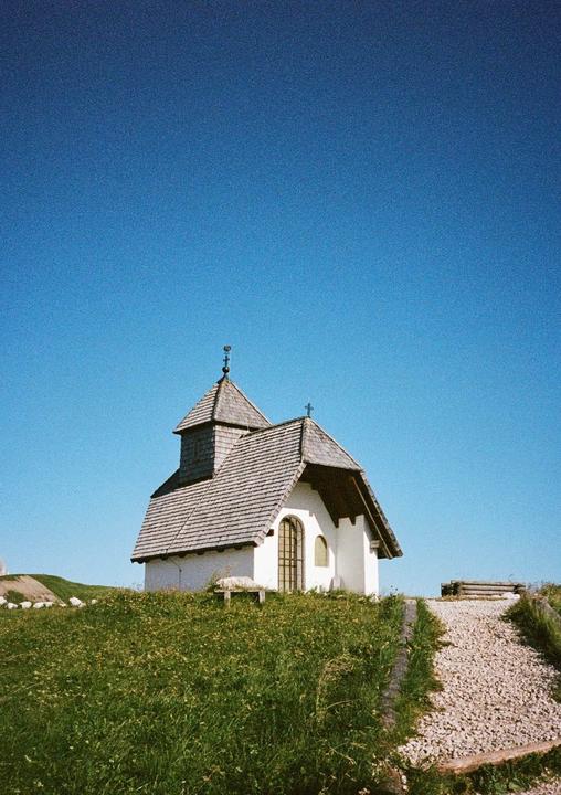 A small white chapel with a shingled roof stands on a grassy hill under a clear blue sky.