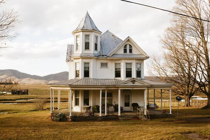A large white Victorian house with a turret and a wrap-around porch stands in a field with mountains in the distance.