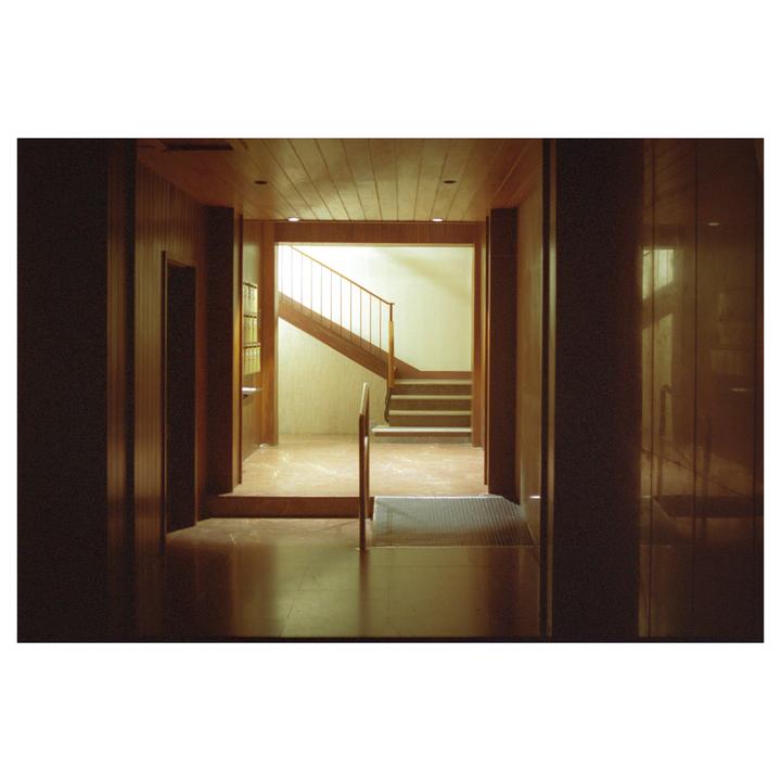 A wooden hallway leads to a brightly lit stairwell with a metal railing.
