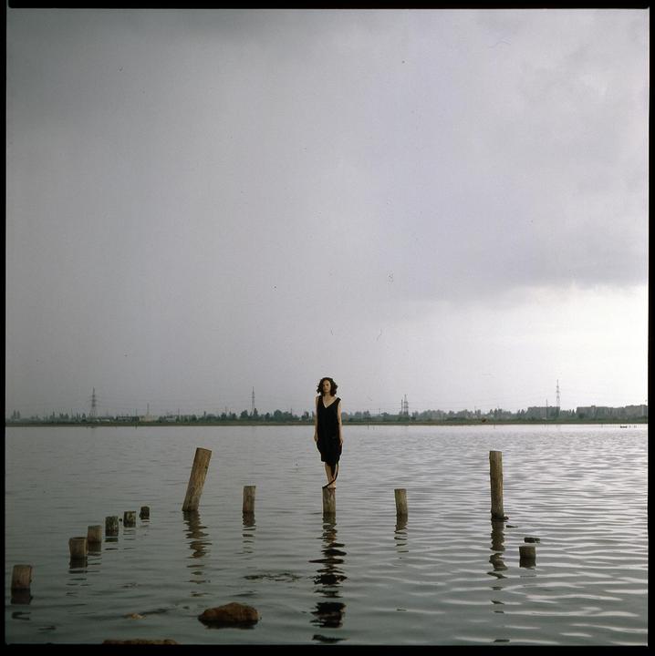 A woman in a black dress stands on a wooden post in the middle of a body of water under a cloudy sky.