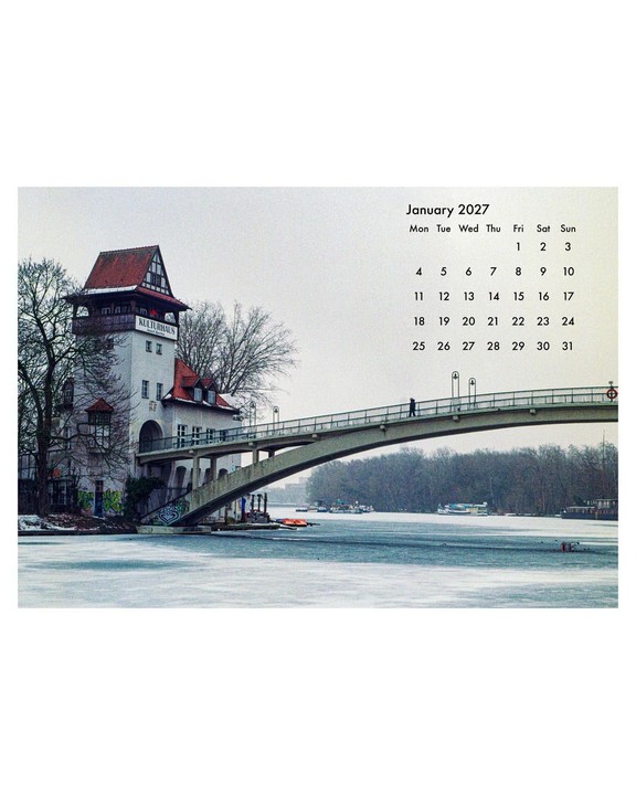 A person walks across a bridge over a frozen river next to a building with a red roof and a tower.