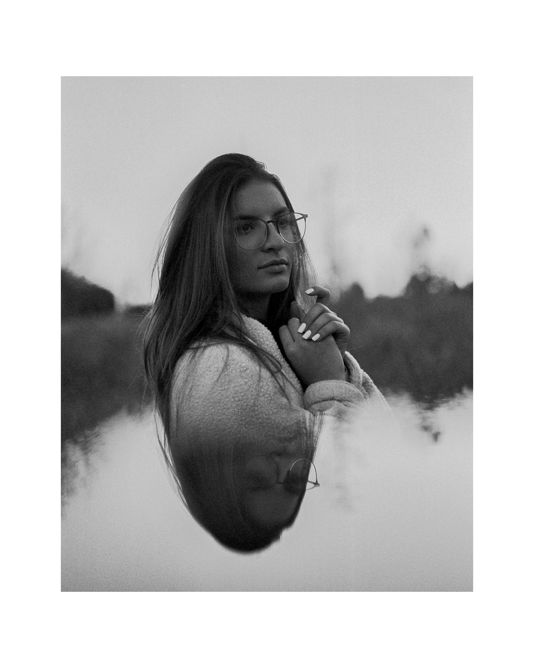 A woman wearing glasses and a fleece jacket is reflected in water, with her hands clasped together.