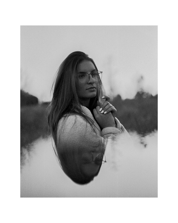 A woman wearing glasses and a fleece jacket is reflected in water, with her hands clasped together.
