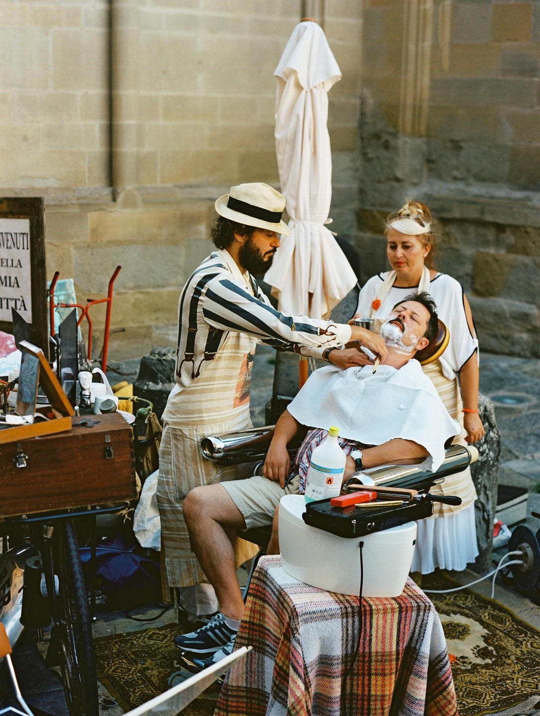 A man in a striped shirt and hat shaves another man's face while a woman watches in a street.