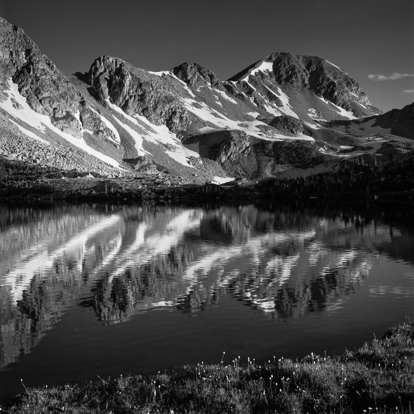 Snow-capped mountains are reflected in a still lake with a grassy shore.