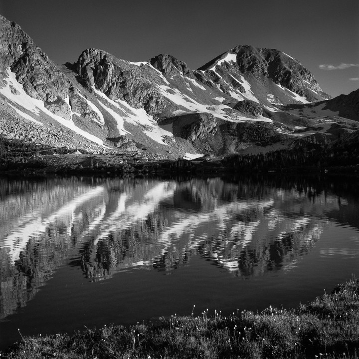 Snow-capped mountains are reflected in a still lake with a grassy shore.