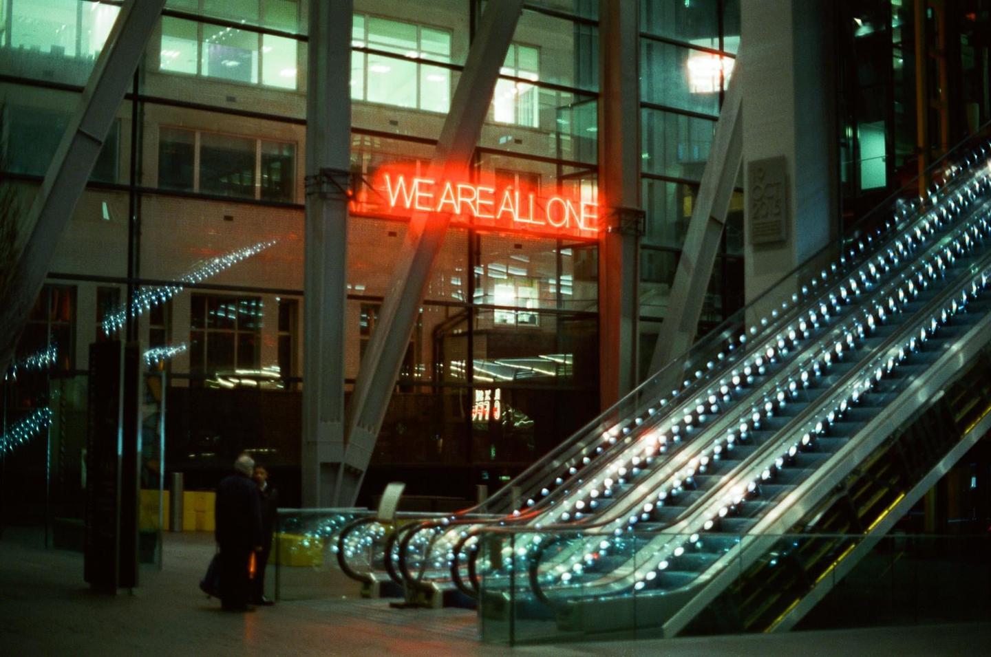 A neon sign reading "WE ARE ALL ONE" hangs above an escalator at night.
