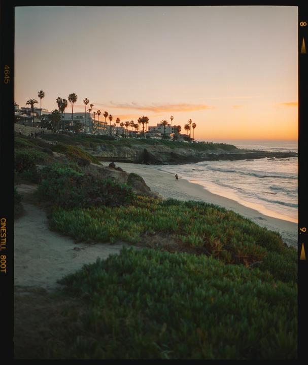 A beach with waves and a person walking on the sand is visible at sunset, with buildings and palm trees on a cliff overlooking the ocean.